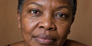 A close-up portrait of a Black woman in her late 50s with bare, unretouched skin showing natural texture, pores and expression lines in soft natural light.