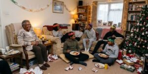 Three generations of a Black family sitting together in a crowded living room, talking and laughing casually on Christmas Day.